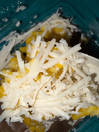 A close-up, top-down shot shows a portion of a teal-colored plastic container filled with yellow corn kernels and shredded white cheese. The corn kernels are visible beneath a thick layer of finely shredded cheese, some of which appears slightly clumped together. A thin, brown, dried strand is present on top of the cheese. The lighting in the image is somewhat harsh, creating highlights and shadows on the food and container. No people, actions, or interactions are depicted. The location is unknown.