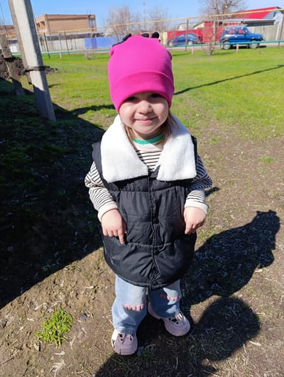 A young child stands on dirt and grass in Уральский, Russia. The child wears a pink hat, a black vest with a white collar over a striped shirt, and blue jeans with pink heart patches on the knees, paired with pink sneakers. The child's shadow falls to the right. In the background, there's a green lawn with scattered leaves, a fence, a blue car, and a small building with a red roof. A utility pole is visible on the left. The weather appears sunny.