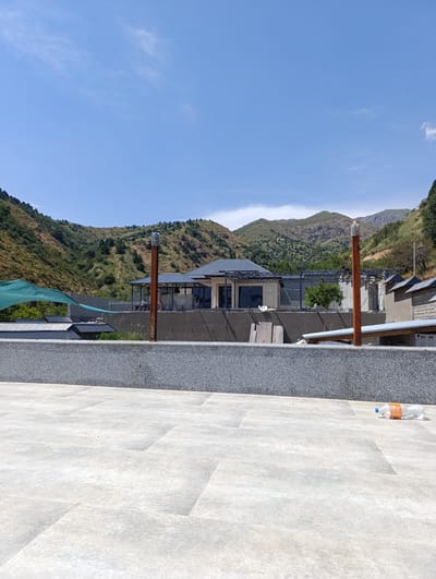 The image captures an outdoor scene on a clear, sunny day. In the foreground, a light-colored tiled patio extends across the lower portion of the frame. To the right on the patio, there is a partially empty clear plastic bottle with an orange label. Behind the patio is a grey stone retaining wall.
Beyond the wall, a construction site is visible. Buildings with dark, angled roofs are under construction, with exposed metal framing in some areas. A green shade cloth is stretched over a section of the site. Several wooden posts, one notably taller and topped with a plastic bottle, stand in front of the construction.
The background is dominated by a mountainous landscape. The slopes are covered with dry, sparse vegetation and scattered trees. The sky above is a clear, light blue with a few wispy white clouds. The overall impression is a semi-arid, mountainous environment with ongoing construction activity. The location is identified as Saylyk, Uzbekistan.