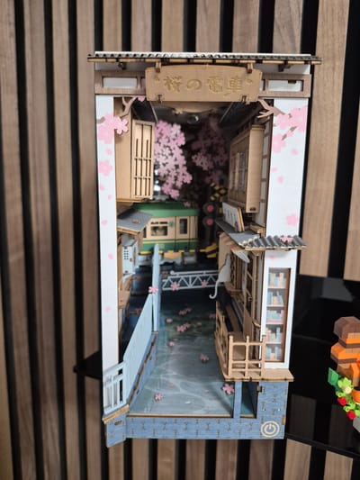 The image shows a diorama of a Japanese street scene with cherry blossoms and a train. The diorama is a detailed model, likely a book nook, featuring traditional Japanese architecture, a train with green carriages, and a railroad crossing with signal lights. Pink cherry blossoms are depicted on the trees and scattered on the ground, creating a serene atmosphere. A white cat with a curved tail is also visible, adding a whimsical touch. The diorama is placed on a black reflective surface, with a vertical wooden slatted background. The overall impression is one of intricate craftsmanship and a charming, idyllic scene. There is no indication of the location being Barbing, Germany; the scene strongly suggests a Japanese setting.