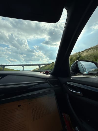 The image captures an interior view of a vehicle's dashboard and windshield, looking out towards a cloudy sky and a distant elevated road or bridge. The foreground is dominated by the dark interior of the car, with a laptop open on the dashboard displaying text. A side-view mirror reflects the road ahead and other vehicles. To the right, a grassy hillside slopes upwards, catching the sunlight. The overall impression is one of travel, with a focus on productivity or work being done while on the move. The specific location of Rupkite, Bulgaria is not visually evident from the provided image.