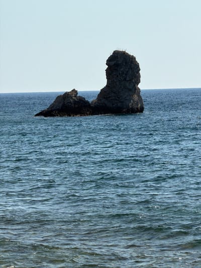 This media file features a close-up, vertically oriented view of a rock formation jutting out from a body of water, likely a waterfall or a strong current. The rock is dark and craggy, with some lighter, possibly dry vegetation clinging to its surface. Behind and to the right of the rock, a continuous flow of water cascades or moves with great force, creating a textured, rippling surface that dominates the right side of the frame. The water appears to be a cool, bluish-gray hue, catching the light in places, suggesting either a bright, overcast sky or the reflective properties of the water itself. To the left of the rock and water is a vast expanse of pale, almost white sky. The overall impression is one of natural elements in motion and repose, with the solid, dark rock contrasting sharply with the fluid, dynamic water and the open, serene sky. The image evokes a sense of the power and texture of nature. No people, text, or specific time of day or weather conditions are discernible beyond the light reflecting off the water. The location context suggests this might be a coastal or mountainous region in Montenegro.