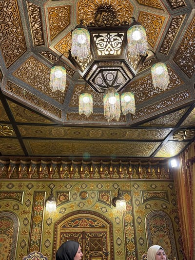 An ornate ceiling displays intricate geometric patterns and floral motifs in shades of gold, olive green, and brown. The ceiling features several chandeliers with a modern design, hanging from elaborate structures. Parts of two women in headscarves are visible on the right side. The overall composition gives off a luxurious and artistic atmosphere.