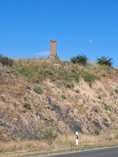 A stone monument, topped with a bird-like carving, stands on a grassy hill under a clear blue sky in Malishka, Armenia. The monument is the central focus, positioned on the crest of the hill. The surrounding environment consists of dry, sparse vegetation and rocky terrain, characteristic of a hillside. A white road marker with a black symbol is visible at the base of the hill, near a road. The scene is static and devoid of human presence.