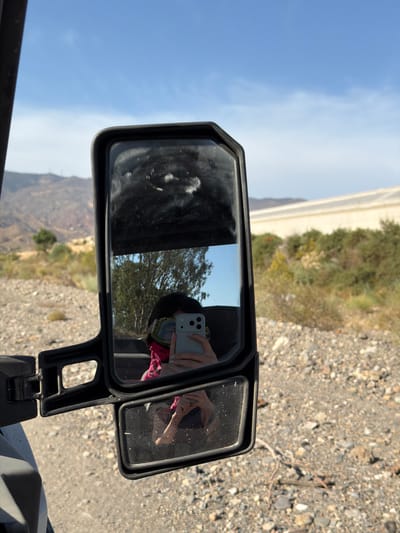 A person is taking a selfie in a side-view mirror of what appears to be an off-road vehicle. The vehicle is parked on a gravel road in a rural area. In the main mirror, a person is visible wearing goggles and a pink scarf, holding a smartphone. The background in the main mirror shows trees and mountains, consistent with the landscape of Pechina, Spain. A smaller, lower mirror reflects the person's arm and hand holding the phone. The environment is dry and arid, with sparse vegetation and a clear blue sky.