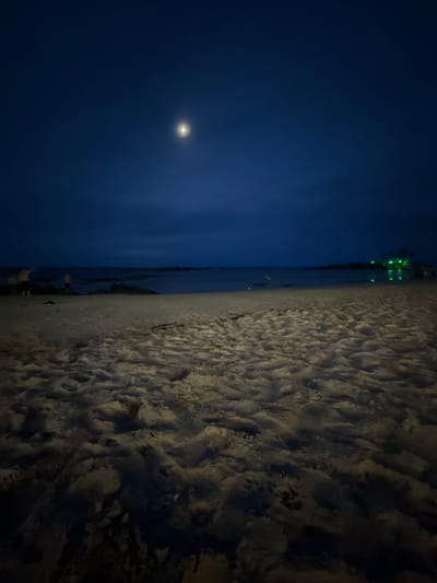 This image displays a nocturnal beach scene in Cairu, Brazil. The sky is a deep indigo, dominated by a bright, circular full moon positioned in the upper-center. Below, a wide expanse of light-colored sand, marked by numerous footprints and undulations, stretches from the foreground to the midground, illuminated by a low-angle light source. The ocean beyond is dark and calm, with a subtle horizon line. On the distant right shoreline, a cluster of vivid green lights indicates structures, likely buildings, whose reflections are faintly visible on the water. On the distant left, indistinct dark formations, possibly rocks or land, are present near the water's edge. Several human figures are discernible on the beach near the water: two indistinct figures on the far left, one with an arm raised; a single figure in the mid-center appearing to be bent over; and a partially visible figure on the far right. The low-light conditions obscure specific details of the individuals and their actions.