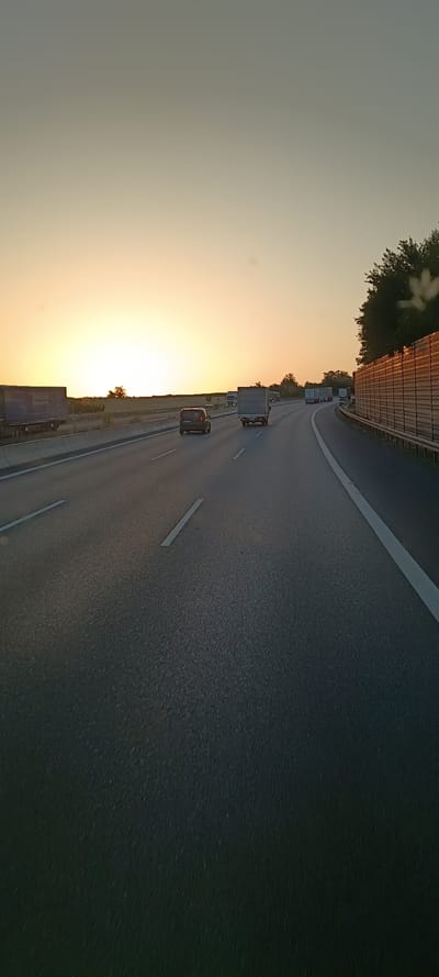 The image depicts a highway scene at sunset in Wiesloch, Germany. The sun is low on the horizon, casting a warm glow across the sky and illuminating a multi-lane asphalt road. Several trucks and a dark colored car are visible traveling on the highway. On the right side of the road, a sound barrier made of wooden panels with metal supports lines the highway. Trees and vegetation are visible in the background. The image appears to have been taken from a moving vehicle.
