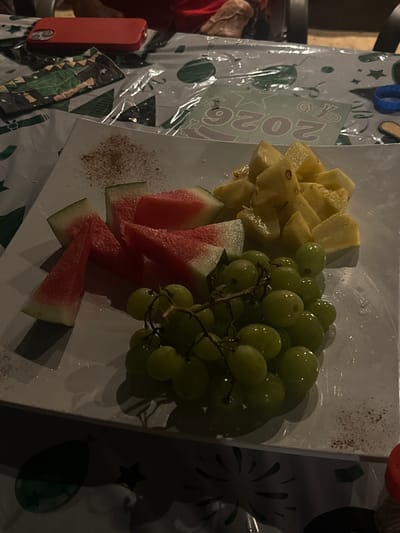 The image shows a plate of fruit containing pineapple, grapes, and watermelon. The plate rests on a festive tablecloth decorated with images of stars, balloons, and the year "2026," along with a graduation cap. To the left of the plate, a red phone is visible. The location is Southlake, United States of America.