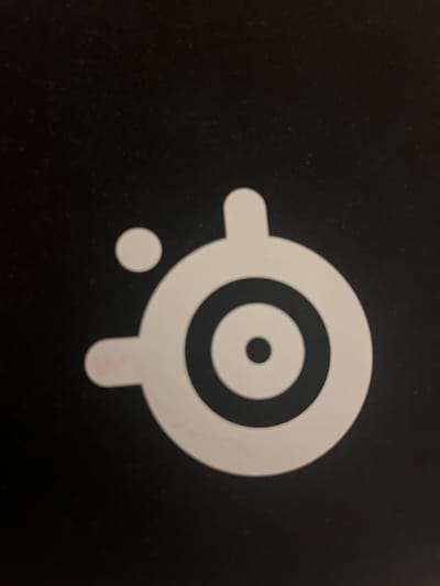 The image displays the SteelSeries logo, which is a white stylized target symbol, set against a black background. The logo consists of concentric circles, with a smaller solid circle in the center and a larger circle surrounding it. Extending from the larger circle are two curved shapes that resemble ear-like protrusions. Below and to the left of the main target symbol is a smaller, solid white circle. The overall impression is a clean, minimalist design. There is no visible text within the image. The lighting is even, suggesting an indoor setting, and there are no environmental cues to indicate the time of day or weather conditions. The image appears to be a photograph of the logo, possibly a sticker or a printed design on a surface. Given the provided location context of Pobiedziska, Poland, this logo might be associated with a local gaming community, an esports event, or a product available in that region.