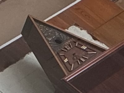 A triangular, wooden clock is angled in the foreground of the image. The clock face is dark with Roman numerals and has two hands. The clock is mounted on a wall with a textured, light brown finish. To the right of the clock, a dark brown, wooden surface is visible. The background consists of a lighter brown textured surface. The image appears to be taken indoors, and there are no people or animals present. The specific location within Melitopol, Ukraine, cannot be determined from the visual information provided.
