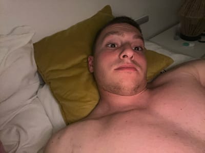 A shirtless man lies in bed, his head resting on a yellow pillow. White bedsheets surround him. He is looking directly at the camera with a slightly furrowed brow. A bedside table is visible on the right, with a lamp and other objects on it. The location is unspecified and there is no evidence to suggest the location is in Vitry-sur-Seine, France.