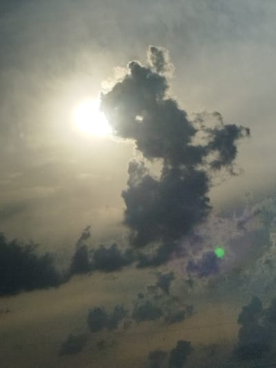 The image is a photograph of a cloudy sky with the sun partially obscured by a dark cloud formation. The sun appears as a bright, blown-out orb in the upper left quadrant of the image. A large, dark, and textured cloud dominates the center and right side of the image, with lighter clouds surrounding it. There is a small, bright green circular lens flare in the lower right quadrant. The overall lighting suggests it is either sunrise or sunset. The image is composed of natural elements in the sky and does not depict any urban or specific geographic features. Therefore, it is not possible to determine if the location is Borcea, Romania, or any other specific city.