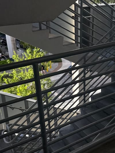 The image presents an outdoor view from an elevated position within a concrete and dark grey metal stairwell structure in Aventura, United States. The stairwell occupies the majority of the frame, featuring multiple flights of grey concrete steps and a complex railing system composed of horizontal and diagonal bars, casting distinct shadows.
Below and beyond the stairwell, a curved, light-colored paved walkway with a brick-like pattern is visible. A male individual, dressed in a dark shirt and light-colored patterned shorts, is walking on this pathway from left to right, holding a white rectangular object in his left hand. The walkway is flanked by several vibrant green trees with dense foliage and multiple yellow cylindrical bollards. To the left of the walkway, an outdoor seating area with tables and umbrellas is partially visible next to a large, light-colored architectural column. Vehicles are faintly discernible in the background. The scene is illuminated by strong natural sunlight, creating defined shadows.
