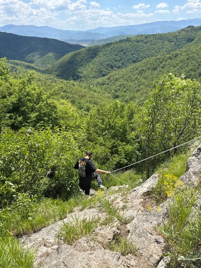 A man is suspended from a cliff face in the vicinity of Skravena, Bulgaria. He is wearing dark clothing, a backpack, and light-colored shoes. He is secured by a rope running diagonally upward towards the rocky terrain at the top of the frame. The cliff edge is covered with grass and some yellow flowers. The background consists of a dense forest landscape and distant mountains under a partly cloudy sky.