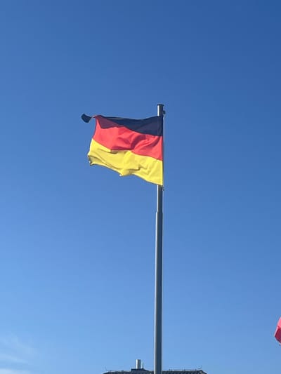 A German flag is depicted waving from a flagpole against a clear blue sky. The flag consists of three horizontal bands of black, red, and yellow. The flagpole is a tall, slender metallic pole, positioned in the lower center of the frame. The upper portion of the flag is taut and sharply creased, while the lower portion billows outwards, creating a dynamic sense of movement. Below the flag and flagpole, a building's roofline is partially visible, suggesting an urban or developed area. The overall scene is dominated by the bright sky and the prominent national symbol. The location is not specified as Niederwald, Germany, but it is presented as a general outdoor scene.