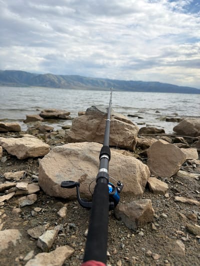 This is a first-person perspective, eye-level shot of a fishing rod resting on a rocky shore by a body of water. The fishing rod is dark gray with a black handle and a blue reel. The foreground is filled with various sized rocks and pebbles. In the background, a vast body of water stretches to a distant, mountainous horizon under a cloudy sky. The location is the shore of Lake Alakol, near the town of Alybai, Kazakhstan.