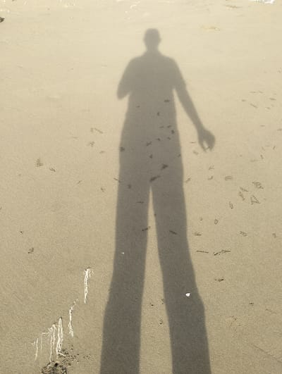A full-body shadow of a person stands cast upon a sandy beach. The shadow is long and elongated, suggesting a low sun angle. The sand is a light brown color and appears damp, with small indentations that could be footprints or crab holes. Some sparse, light-colored debris, possibly seaweed or small shells, is scattered on the sand. The shadow shows the outline of a head, torso, arms, and legs. One arm is extended to the side. The background is out of focus, but appears to be more of the sandy beach and possibly a body of water. The location is identified as Blanglancang, Indonesia.