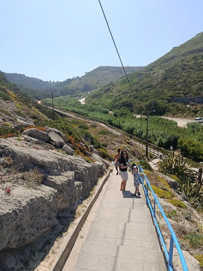 This is a full-shot, eye-level photograph taken on a sunny day. In the foreground, a concrete staircase with a bright blue metal railing descends. Three people are visible on the stairs. On the left, a woman in a black t-shirt and light-colored shorts stands facing the camera, holding a phone. Next to her, a young boy wearing a patterned t-shirt, shorts, and a baseball cap, leans on the blue railing. Behind them, a man with dark hair and wearing dark shorts walks away from the camera.
The staircase is bordered by rocky terrain on the left, with scattered green and orange vegetation. On the right, the railing continues alongside a slope covered in low-lying green and yellow plants.
The middle ground shows a wooden boardwalk winding up a grassy slope, with a few scattered trees. Beyond the boardwalk, a dirt road is visible, leading towards a cluster of green vegetation. A white vehicle is parked on the right side of the road.
The background features rolling, green hills under a clear blue sky. A power line extends diagonally from the upper left corner of the image. The scene is set in the natural landscape near São João das Lampas, Portugal, likely a hiking or tourist area.