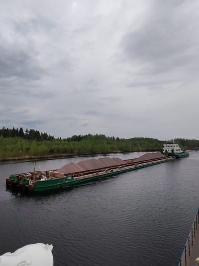 The image shows a barge being towed on a waterway, likely a canal, in an area with overcast skies. A long, green barge filled with what appears to be gravel or sand is being towed by a white and green tugboat. The barge is marked with the number "7703". The scene is set on a body of water with rippled surface. Trees line the edge of the waterway on one side. The photo is taken from a slightly elevated perspective, potentially from another vessel or structure near the water. It is most likely taken in the vicinity of Alekseevskoe, Russia.