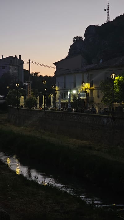 The image depicts an outdoor scene at dusk in Alhama de Aragón, Spain, featuring a waterfront restaurant and a body of water. A stone wall runs along the edge of a gently flowing body of water, likely a river or canal, with a grassy bank on the left. On the right side of the water, a terraced area in front of a building is set up for outdoor dining. Several tables with people seated are visible, with white umbrellas providing shade. Lampposts illuminate the area. The building behind the terrace has multiple windows and a sign that reads "EL CLÁSICO." To the left, a vertical sign for "TITO RESTAURANT" is visible. In the background, a rocky hillside dominates the skyline, topped with a telecommunications tower. Residential buildings are also present on the left side of the image. The sky is a gradient of soft purple and orange, indicating the transition from day to night.