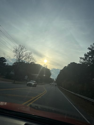 The photograph captures a view from a vehicle, looking down a sunlit road. The road has double yellow lines and cars are present. Trees line both sides, casting shadows. The sun, low in the sky, creates a glare. Power lines run parallel to the road on the left. The sky is mostly cloudy with some clear patches. The image is slightly rotated.