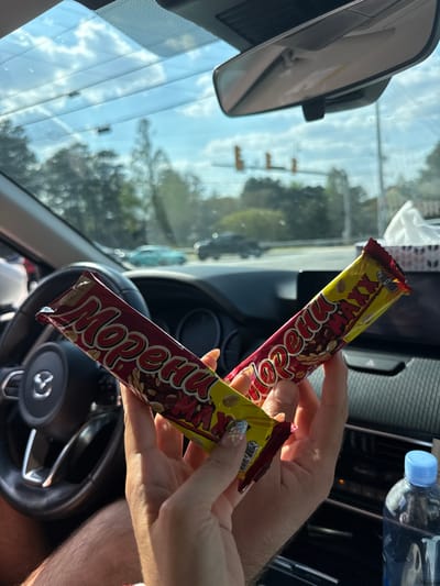 The scene is captured from inside a Mazda vehicle, stationary on a road in Marietta, United States. Two hands are centered in the foreground, holding two identically packaged "Морени MAXX" candy bars. The left hand displays manicured nails (blue with white floral accents on the thumb, yellow on the index finger), while the right hand exhibits visible arm hair. A black Mazda steering wheel is visible to the left. A blue-capped plastic water bottle and a white tissue box are present on the right-side dashboard. Through the front windshield, a daytime sky with scattered clouds, blurred green foliage, and traffic lights are visible. A light blue vehicle and other blurred traffic are present on the road ahead.