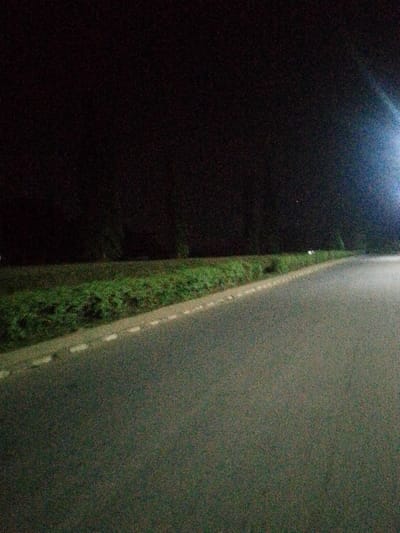 A dark, grainy image shows an empty asphalt road at night. The road curves slightly to the right. A narrow strip of green vegetation lines the left side, separated from the road by a white-dashed curb. The sky above the vegetation is black. A bright light source is faintly visible in the upper right, creating a glow.