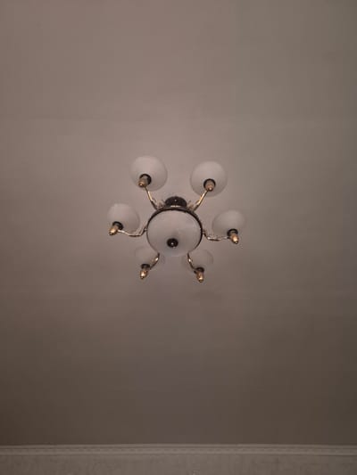 A vintage, multi-armed chandelier is centered against a plain, light-colored ceiling. The chandelier features a central globe-shaped fixture and six smaller, round glass shades, each supported by a gold-toned arm with decorative accents. The ceiling appears to be textured. The image is a vertical shot taken from below, looking upwards. There is a subtle horizontal line of what appears to be decorative molding or trim visible at the very bottom of the frame. There are no people or animals present. The lighting is subdued. The scene is indoors, in what could be a residential setting. The city of Қанабек, Kazakhstan is not depicted in this image.