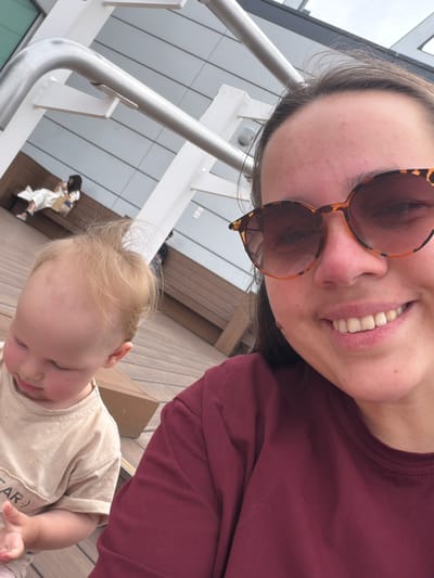 A medium shot captures a woman with sunglasses and a baby on a wooden deck, possibly outdoors given the light-colored building in the background. The woman, in the foreground on the right, wears a maroon shirt and tortoiseshell sunglasses, smiling at the camera. Her hair is dark brown. The baby, to the left, wears a light-colored t-shirt and is looking down. The background shows a woman in a white outfit sitting on a bench and a man walking in the distance. The scene appears to be in the city of Ota, Japan, likely at a public space or a commercial area with an outdoor seating section.