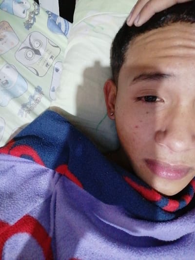 This image depicts a male individual lying in bed, likely indoors, within Soacha ciudad, Colombia. The subject is seen from the chest up, positioned at an upward angle, looking directly towards the camera. The right side of his face is partially shadowed. His right hand is resting on the top of his head/forehead, with fingers visible. He has short, dark hair and a light complexion. A small, spherical earring is visible in his left earlobe. He is partially covered by what appears to be a purple fleece blanket or garment, which has an underlying blue pattern with red circular elements. The pillow beneath his head is light green with cartoon-style animal patterns, including an owl and bears. The background suggests a domestic bedroom setting.