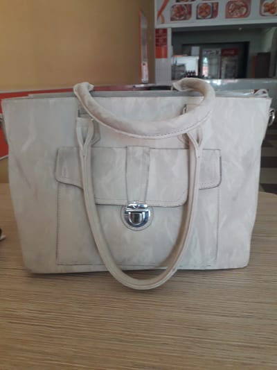 The image features a light-colored handbag, possibly off-white or cream, resting on a wooden table or counter. The bag is oriented vertically, extending across the frame, and displays a subtle textured or patterned surface. It has a single handle and a front flap secured by a silver-toned metallic clasp. Details like stitching along the flap and straps are visible, and there are slight signs of wear and discoloration on the bag, particularly on the handle and around the clasp area.

The setting appears to be an indoor commercial space, likely a cafe, restaurant, or food establishment. In the background to the left, a vertical display of framed pictures showcases various food dishes, suggesting a menu or promotional board. Below these food images, red and white structural elements are visible, possibly part of a counter or seating area. Further into the background, through an opening, blurred objects resembling kitchen equipment, shelves, and potentially a beverage refrigerator with faint red branding are discernible. The wooden surface beneath the bag extends to the right, exhibiting a distinct vertical wood grain pattern.

No individuals are present, and no specific activity is occurring. The time of day and weather conditions are indeterminate due to the indoor lighting. There is no clearly legible text visible in the image. The overall impression is that of a bag casually placed on a table within a public dining or service venue.