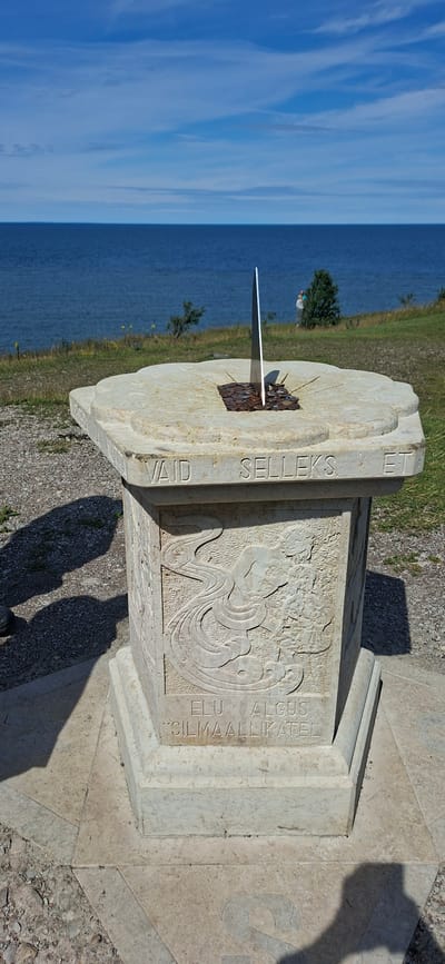 The image depicts a sundial monument in Panga, Estonia. The sundial is made of light-colored stone and features an engraved base with decorative carvings and text. The top surface of the sundial has a metal gnomon and is filled with numerous small coins. The monument is situated outdoors, with a backdrop of the blue sea and sky, and a grassy slope. In the distance, two figures can be seen near a tree, and there are shadows cast on the ground from the monument and possibly other unseen objects. The ground around the monument is covered with gravel and stone. The carved text on the base includes "VAID SELLEKS ET" and "ELU ALGUS SILMAALLIKATEL".
