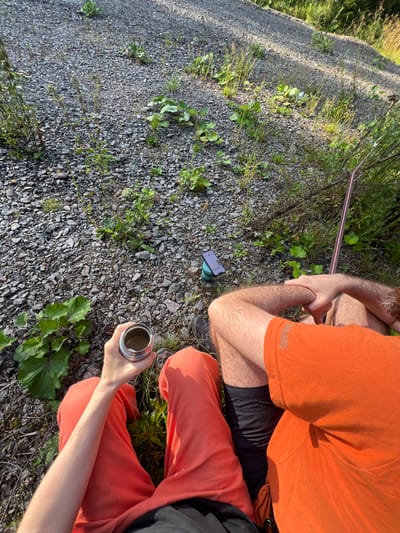 The image shows two individuals sitting outdoors on a gravelly surface with sparse vegetation, likely in a natural or semi-natural area. The setting appears to be a pathway or embankment, with the ground covered in small stones and occasional green plants. One person is wearing an orange t-shirt and black shorts, while the other is wearing orange pants.

In the foreground, one individual's hand holds a silver metallic thermos or mug. In the mid-ground, a smartphone is propped up on a teal-colored stand. A thin pink and black strap, possibly part of a bag or equipment, extends diagonally across the upper right portion of the image. The lighting suggests it is daytime, with sunlight casting shadows.

Without further information, it is not possible to definitively place this scene within the city of Hämeenlinna, Finland. The environment shown is generic and could be found in many locations.