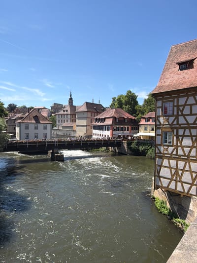 The image captures a sunny day scene in Bamberg, Germany, focusing on the Regnitz River flowing through the historic city. In the foreground, the river's surface is textured with ripples and reflections of sunlight. A pedestrian bridge spans the river, adorned with flower boxes, and several people are walking across it.
On the far side of the river, a cluster of traditional half-timbered buildings lines the bank. These buildings exhibit characteristic architectural styles of the region, with exposed wooden beams and plaster infill. To the right of the bridge, a prominent half-timbered building juts into the frame, its facade featuring a distinctive pattern of wooden supports and light-colored infill. Its tiled roof has a dormer window with a small pane.
Beyond the bridge and the immediate riverfront, more buildings of varying architectural styles are visible, including some with pitched roofs and shuttered windows. A church tower with a spire rises above the buildings in the background. Lush green trees partially obscure some of the structures, indicating a verdant environment. The sky above is a clear, bright blue, with a faint contrail visible high up. The overall impression is one of a picturesque and lively urban landscape on a pleasant day.