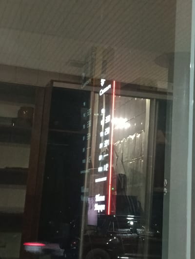 The image shows a reflection in a window at night. The reflection features a vertical neon sign with white text that appears to be a price list or advertisement. The text is difficult to read clearly due to the reflection and low light conditions, but some numbers are visible, such as "320", "238", "334", and "12". Above these numbers, a logo or word that looks like "Coron" is partially visible. Below the numbers, there is text that reads "Supermarket". The scene also reflects a dark car and possibly people inside. The overall atmosphere is dark, suggesting it's nighttime, and the location appears to be a commercial area, likely a supermarket or a similar establishment in Besagash, Kazakhstan. The reflections create a layered effect, making it challenging to distinguish between the actual scene and its mirrored image.