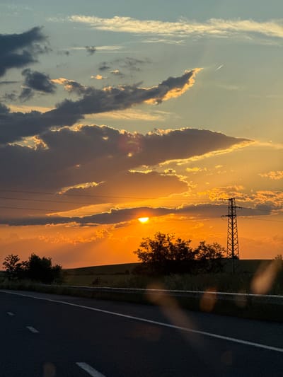 A photograph depicts a vibrant sunset, partially obscured by dark clouds, with beams of light breaking through. A utility pole with wires is visible near the center of the frame. Trees are silhouetted against the bright sky. A portion of an asphalt road with white lines runs along the bottom right of the image. There is no evidence to confirm this picture was taken in the city of Zetiovo, Bulgaria. No people are visible in the frame.