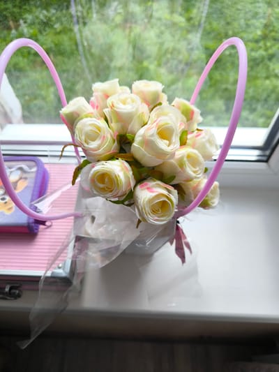 A bouquet of artificial white roses with pink tips is displayed in a white container with a pink ribbon. The container is placed on a white windowsill, with a window behind it looking out onto greenery. To the left of the roses is a small pink zippered pouch with a cartoon character on it. The scene appears to be indoors, possibly in a home or room, with natural light coming from the window. The overall impression is one of simple decoration or a gift arrangement.