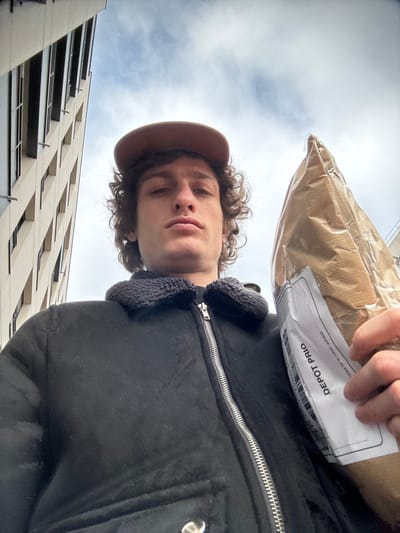 A low-angle selfie captured in Joinville-le-Pont, France, depicts a male individual from the chest up, positioned against a backdrop of urban architecture and sky. The individual has medium-length curly brown hair and wears a light brown baseball cap. He is dressed in a black jacket featuring a dark, possibly fleece, collar and a prominent silver zipper running down the front. He looks directly at the camera with a neutral expression. In his left hand, on the right side of the frame, he holds a package wrapped in light brown paper and clear plastic. A white shipping label, vertically oriented, is affixed to the package, displaying a barcode and the text "DEPOT PRIO." The background on the left features a portion of a tall, modern building with light-colored facade panels and multiple rectangular windows. Above the building and the subject, a partly cloudy sky is visible.