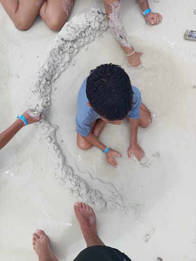 In an overhead shot, several individuals are interacting with a sandy, wet environment, likely a beach or a water-filled sandy area. The sand appears light-colored, almost white, and is mixed with water, creating shallow pools and a slick surface.
In the foreground, a pair of bare feet is visible, appearing to stand in the shallow water and sand. Further back, a young person wearing a blue shirt is crouched down, with their hands in the sand, appearing to be playing or building something. This individual has dark hair and is wearing a blue wristband.
To the left, another person's arms are visible, also interacting with the sand. One arm has a blue wristband, and the other has a pink wristband. This person's arm is covered in white residue, possibly from the sand.
In the upper left quadrant of the image, the knees and legs of another individual are visible, also covered in the white sand residue. The overall scene suggests a recreational activity taking place in a sandy, wet area. There are no discernible elements that indicate the specific location within the city of Unknown, Colombia.