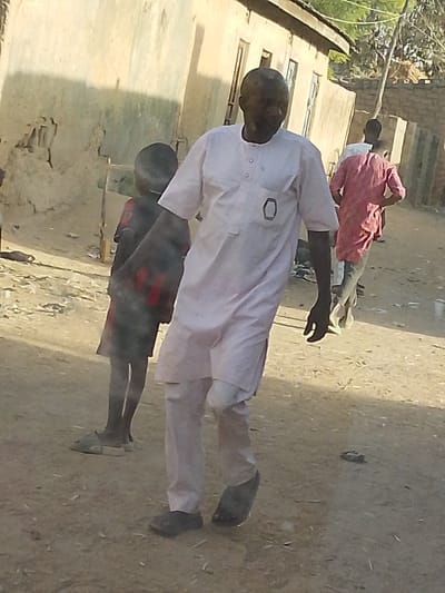 An older man walks on a dusty street, wearing a light pink, long-sleeved traditional outfit and dark shoes. He walks past a child in a patterned shirt. Behind him, another person in a reddish shirt walks further away. Buildings, likely residences, line the street, with one close by displaying weathered, sun-faded walls. The scene appears to be set in a rural or less developed area.