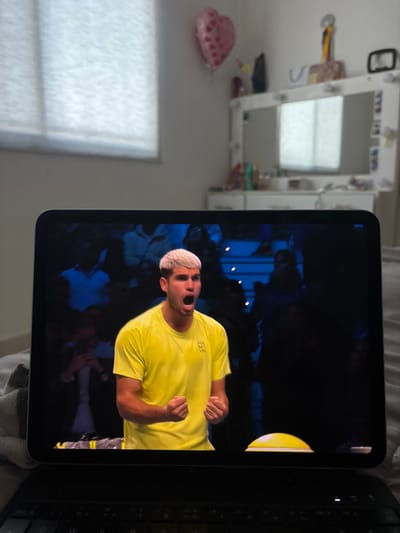 An iPad screen dominates the view, displaying a tennis match scene. A player in a bright yellow shirt, blonde hair, and fair skin is captured mid-celebration, his expression animated. The background reveals a crowd, suggesting a live game. To the right of the iPad is a dark-colored keyboard. In the room's background, a vanity table with a mirror is visible, along with a window covered by blinds and a heart-shaped balloon on the wall.