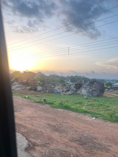 The image depicts a rural or semi-rural landscape in Ribina East, Nigeria, at either sunrise or sunset, viewed from a moving vehicle. The sky occupies the upper half, displaying a bright orange-yellow glow from the low sun on the far left, transitioning to light blue and grey with scattered clouds. Several horizontal power lines traverse the sky. A dark, vertical obstruction, consistent with a vehicle's interior frame, is visible on the extreme left.

The mid-ground features a landscape characterized by numerous large, dark grey boulders scattered amidst green vegetation. Clusters of trees are visible, with denser foliage further in the background. A small, light-colored building is present among the rocks in the mid-left background. On the right side of the background, several distant buildings with light-colored roofs indicate a settlement.

The foreground is dominated by a reddish-brown dirt road that runs diagonally from the lower left to the lower right, appearing somewhat uneven. A small patch of grey, possibly paved, surface is visible on the far lower-left corner of the road. Two dark fowl (possibly chickens) are visible on the dirt road in the mid-left foreground. Scattered pieces of light-colored litter are visible on the road and in the grassy areas. A dark, circular object, possibly a discarded tire or a hanging decorative item, is seen in the grassy area on the mid-right. No people are visible.