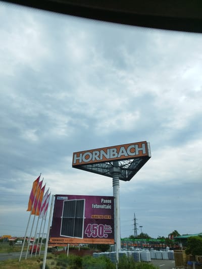 A large, triangular HORNBACH sign is mounted on a metal pole against a cloudy sky. Below the sign, a billboard advertises "Panou fotovoltaic" (photovoltaic panel) for 450 lei, with a "TrinaSolar" logo and an image of two solar panels. Several colorful flags, orange, pink, and white, are displayed on poles to the left of the billboard. In the background, a road with a red car is visible, along with other buildings and a power line tower. The scene appears to be in an outdoor commercial area, possibly a parking lot or storefront, with stacks of goods and greenery. The location is identified as Domnești, Romania.
