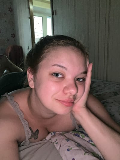 This media file features a young woman lying in bed, taking a selfie. She is positioned on her side, with her head tilted and her left hand resting on her forehead. Her eyes are open and looking towards the camera, and she has a neutral expression. She is wearing a light-colored top with lace trim. A tattoo of a butterfly is visible on her left shoulder.
The setting appears to be a bedroom. Behind her, the wall has a horizontal striped pattern. To her left, a window is visible, reflecting light. The overall lighting suggests it might be late evening or early morning. The scene is intimate and personal, with the subject in a relaxed posture. No text is visible in the image. The location context is Gorodets, Russia.