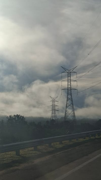 The image depicts two electricity pylons standing tall amidst a misty or foggy landscape, with a dense forest or treeline in the background. The pylons are intricately structured, with crisscrossing metal beams supporting the high-voltage power lines that stretch across the frame. The sky above is overcast, with diffused light suggesting either early morning or a cloudy day. In the foreground, a road or pathway is visible, separated from the natural environment by a metal guardrail. The overall atmosphere is serene and slightly dramatic due to the mist and the imposing presence of the pylons. The scene is located in Lamtamot, Indonesia.