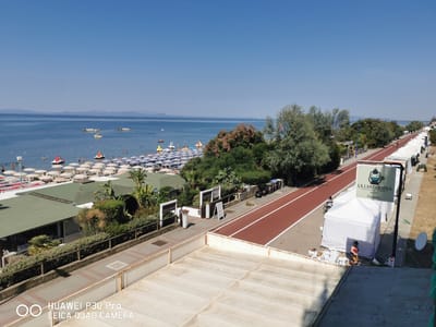 The image shows a seaside view of a beach in Follonica, Italy, on a clear, sunny day. In the foreground, a section of a building's rooftop is visible, with a white railing. Beyond this, a paved walkway with a wooden railing separates the building from a green area with trees and bushes. Further back, a row of white tents and a red running track are present. Several bicycles are parked near the tents. A sign for "LA LANTERNA RISTORANTE PIZZERIA dal 1967 Damoka" is attached to a pole. A woman in a red top and patterned pants is near the tents, and a person on a bicycle is on the running track. The middle ground features more green vegetation, including palm trees and bushes, adjacent to several buildings with green roofs. A wide stretch of beach is covered with numerous striped umbrellas and sunbeds. People are visible on the beach and in the water, with several paddle boats and other watercraft in the sea. The background shows the calm blue sea extending to the horizon, with faint landmasses visible in the distance under a clear blue sky. The overall scene depicts a lively tourist area.