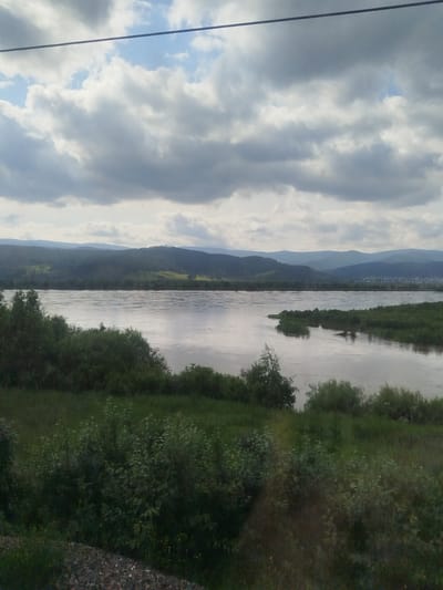 This media file is a landscape photograph taken from a high vantage point overlooking a wide, calm river. The river occupies the central portion of the image, its surface reflecting the overcast sky and the distant, rolling hills. The water appears tranquil, with gentle ripples suggesting a slow current.

The foreground is dominated by lush green vegetation, including bushes and grasses, suggesting a natural, undeveloped area. Some patches of gravel are visible in the lower left corner, possibly indicating a path or the edge of a railway line. Beyond the river, the landscape rises into a series of layered hills, shrouded in a soft, hazy atmosphere. The hills are covered in dense green foliage, with lighter green patches that might be meadows or clearings. In the far distance, a faint hint of a settlement or town can be discerned on the horizon, nestled among the hills.

The sky is filled with dramatic, heavy clouds, ranging in color from bright white to dark grey. The overall lighting suggests it is daytime, likely mid-morning or afternoon, under cloudy conditions. The atmosphere is serene and somewhat muted due to the overcast weather. There are no people or animals visible in the frame. A thin, dark horizontal line stretches across the top of the image, which could be a power line or a part of the structure from which the photograph was taken. The location context of Solnechnyy, Russia, suggests a natural, possibly rural or wilderness setting.