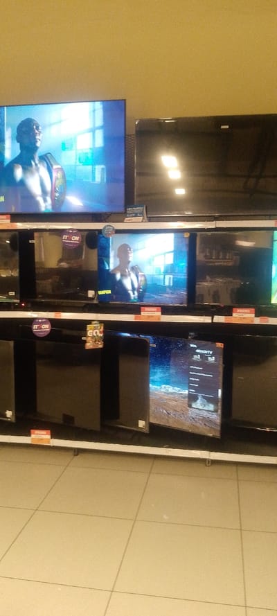 The image displays a retail environment, specifically an electronics section, featuring multiple flat-screen televisions arranged on three horizontal shelves. The walls are uniformly beige, and the floor consists of light-colored square tiles. Overhead lighting is visible through reflections on the screens of the upper televisions.

On the top shelf, a large television on the left is actively displaying content: a shirtless male figure, possibly a boxer, wearing a championship belt and looking upwards. The background behind him appears to be an indoor setting with windows. To the right of this, another large television is powered off or displaying a dark image, reflecting the overhead lights as two distinct square shapes.

The middle shelf presents several televisions. One on the far left is dark. The adjacent television displays the identical image of the shirtless male figure with the championship belt. To its right, another television is off or dark, showing prominent reflections of the environment. Several small, circular purple stickers bearing the text "IT'S ON" are affixed to the televisions or the shelf above them. Below some televisions, red rectangular labels marked "PROMO" are visible.

The bottom shelf also holds multiple televisions. Most of these are dark. One television towards the center is powered on, displaying what appears to be a landscape scene, possibly a desert or barren terrain under a blue sky, with some on-screen interface elements, including "TCL 4K UHD" branding and text. A small, multi-colored promotional tag labeled "GO TV" hangs in front of one of the dark televisions on this shelf.

No individuals are directly visible within the captured scene, apart from the depiction on the active television screens. The specific location within Majengo ward, Kenya, cannot be confirmed from the visual information present in the image.