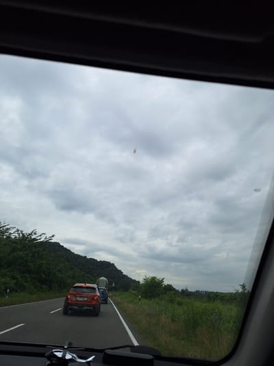 The image captures a view from inside a vehicle traveling on a road through a rural, possibly mountainous, area. The sky is overcast with heavy clouds. In the foreground, the dashboard of the vehicle is visible, with a chrome-plated helicopter figurine mounted on it. The road ahead is paved and has white lane markings. There are several vehicles on the road, including an orange Lada SUV in the foreground, a blue car behind it, and a large truck further ahead. The landscape on either side of the road is lush and green, with dense trees and vegetation, and a hill covered in trees is visible in the background to the left. The overall impression is of a journey through a natural, somewhat unpopulated area on a cloudy day. The location is identified as ნიგოითის თემი, Georgia, which is a community in the western part of the country.