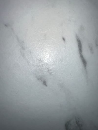 This media file is a close-up, overhead shot of a white marble surface with grey veining. The lighting creates a bright glare in the center of the frame, obscuring some of the detail in that area. The marble has a smooth, slightly textured appearance. There are no people, objects, or identifiable scene elements present. The image focuses solely on the texture and pattern of the marble. The time of day, weather, location cues, emotions, and any visible text are not discernible from the image.